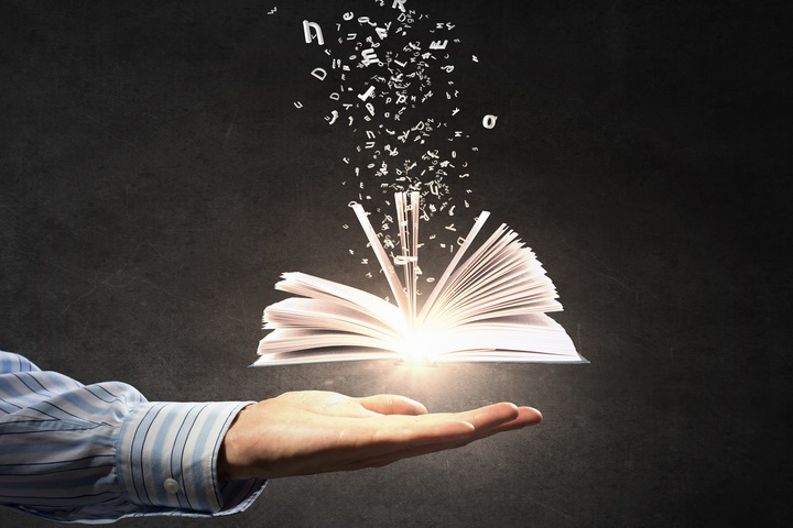 A man wearing a blue shirt holds up a book with magical words coming out of a book.